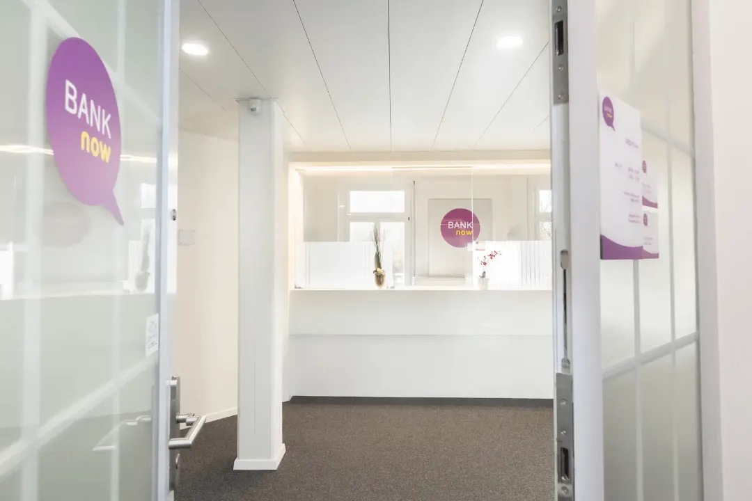 Área de atendimento clara da filial BANK-now Lucerna com balcão de recepção e parede de vidro.