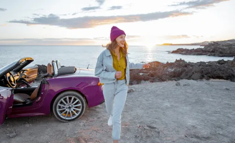 Frau mit pinkigem Cabriolet steht auf einem Parkplatz mit dem Meer im Hintergrund